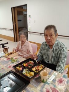 梅雨空に負けない笑顔！地元の特製お弁当で心も晴れやかに うっとうしい梅雨空が続く今日この頃、皆様いかがお過ごしでしょうか。 第二亀岡園では、ジメジメした気分を吹き飛ばすような、心温まる行事食の時間を設けました。じめじめした日が続きますが、皆様とてもお元気にお食事されています。 今回ご用意したのは、地元亀岡で長年愛されている「宮本屋」さんの特製お弁当です。色とりどりのお寿司にてんぷら、彩り豊かなちらし寿司など、箱を開けた瞬間「わぁ！」と歓声が上がるほど豪華な内容。外は雨でも、食卓はパッと明るく華やぎます。 中でも、ひときわ目を引いたのは、仲睦まじくお食事をされている櫟原様ご夫婦のお姿です。 「美味しいなぁ」「こっちのも美味しいで」と、にこやかにお話しされながら、お二人だけの特別な時間を楽しまれているご様子。美味しいお弁当を前に、自然と笑みがこぼれます。 普段の食事も大切ですが、こうして時折、地元の名店の味を皆で楽しむ時間は、入居者の皆様にとって大きな喜びと生活の張り合いになっています。ご夫婦水入らずで過ごす穏やかな時間は、私たち職員にとっても心温まる光景でした。 美味しいお弁当で笑顔を届けてくださった宮本屋さん、本当にありがとうございました。 これからも第二亀岡園では、皆様に喜んでいただけるような楽しい企画を行ってまいります。
